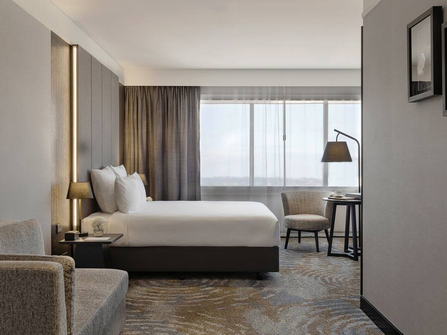A hotel room with a large bed, a side table with a lamp, a chair, a small table, and a view of the city through large windows.