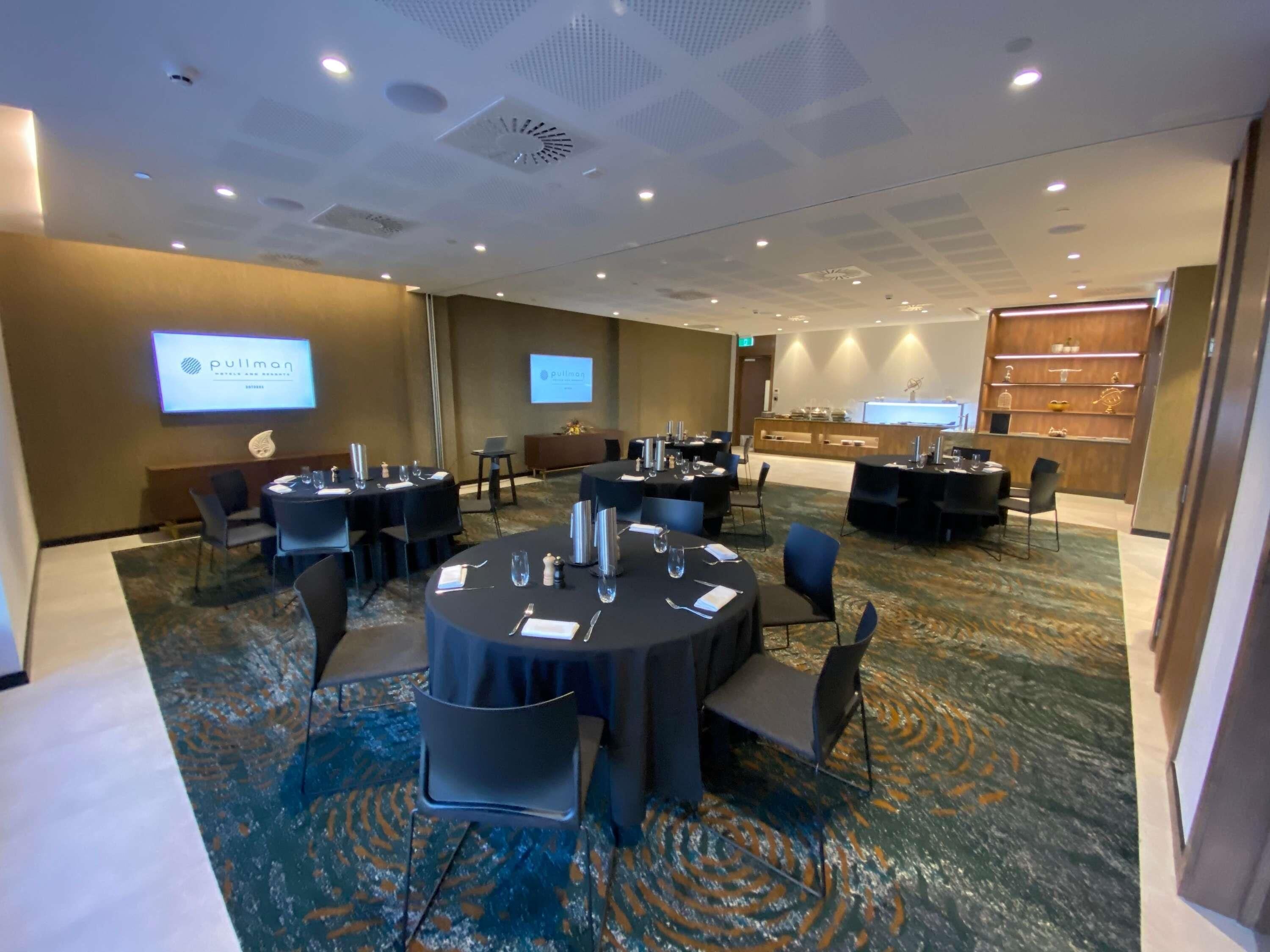 A conference room with round tables, chairs, a projection screen, and a buffet area.