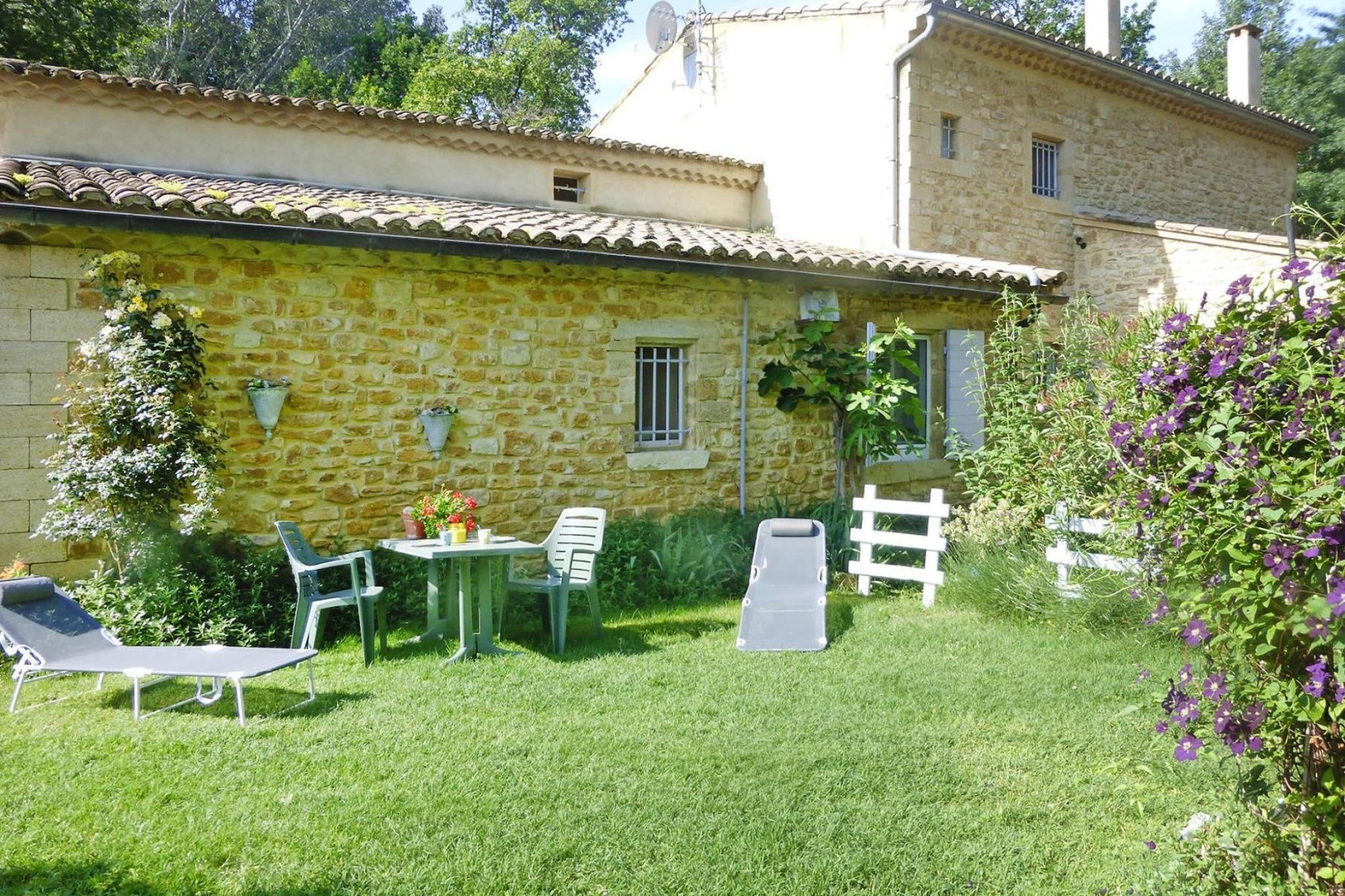 A stone building with a tiled roof, a small garden with a lawn, a table with chairs, and a white fence.