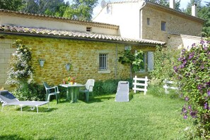 A stone building with a tiled roof, a small garden with a lawn, a table with chairs, and a white fence.