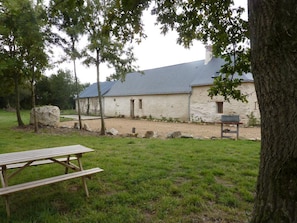Ein rustikales Steinhaus mit geneigtem Dach, umgeben von einer Grünfläche mit Bäumen und einem Picknicktisch.