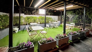 An outdoor patio area with tables and chairs, surrounded by planters with flowers, under a wooden pergola with lighting. - Pensión Cassandra (Arona)