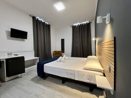 Habitación de hotel con cama, mesita de noche, un escritorio pequeño con computadora, televisor montado en la pared y una lámpara moderna.