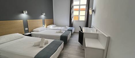 Hotellihuone, jossa on kolme sänkyä, työpöytä ja ikkuna, jossa on verhot.