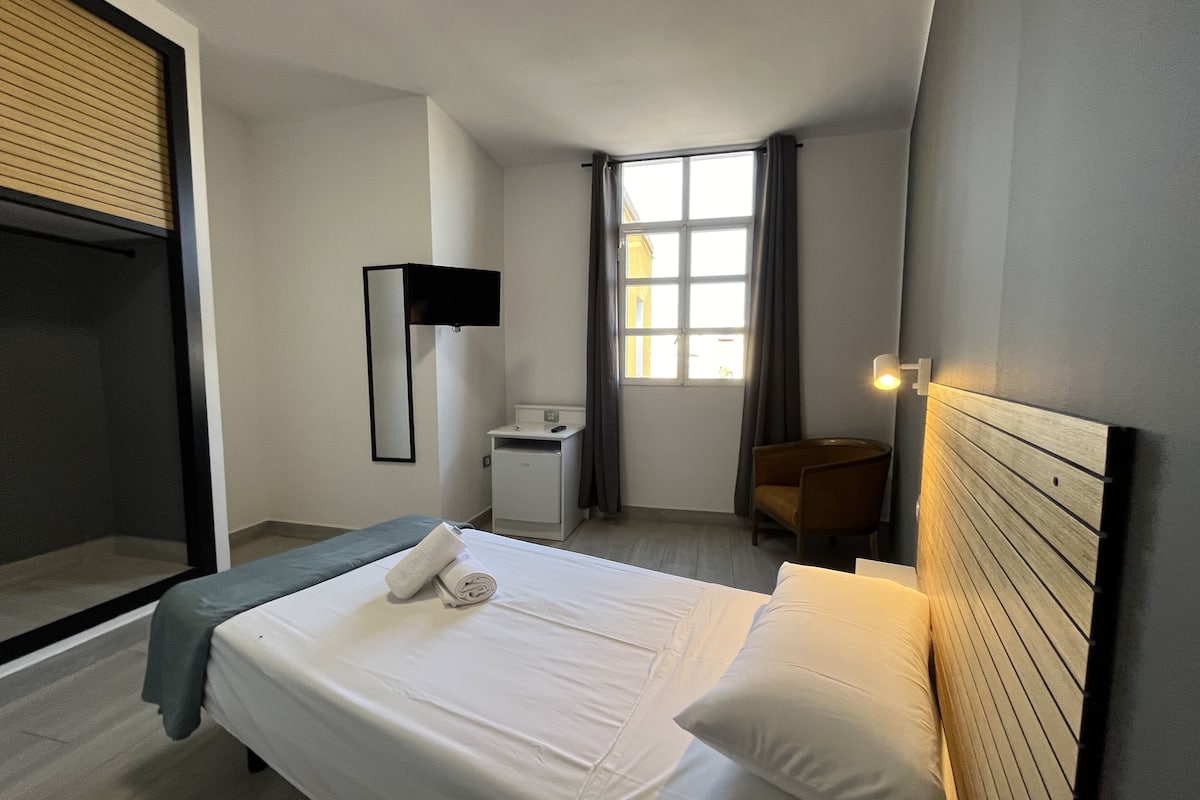 A hotel room with a bed, a desk, a chair, a mirror, a window with curtains, and a nightstand.