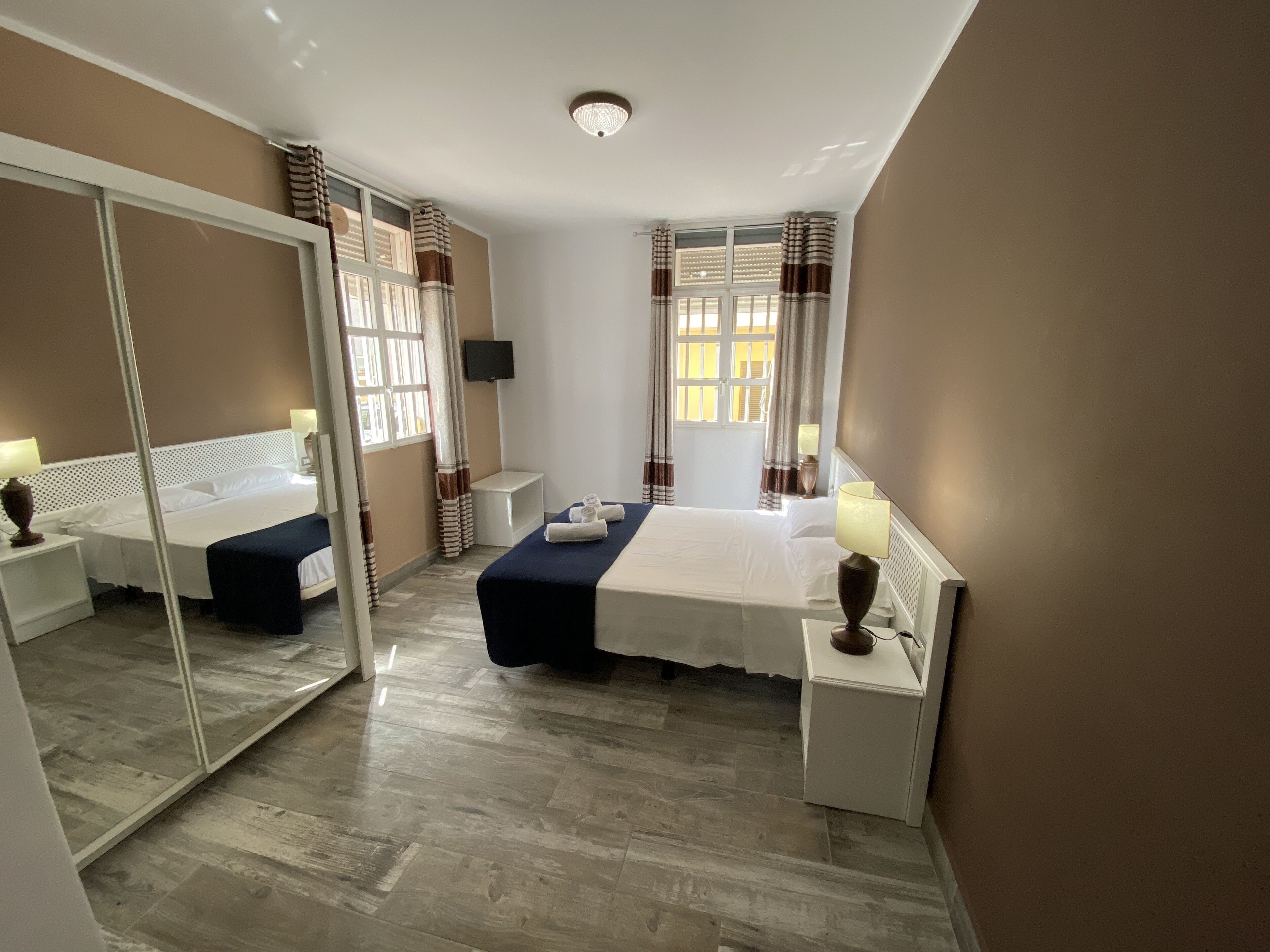 Una habitación de hotel con un espejo grande, una cama con cubrecama azul, una mesita de noche con lámpara y una ventana con cortinas.