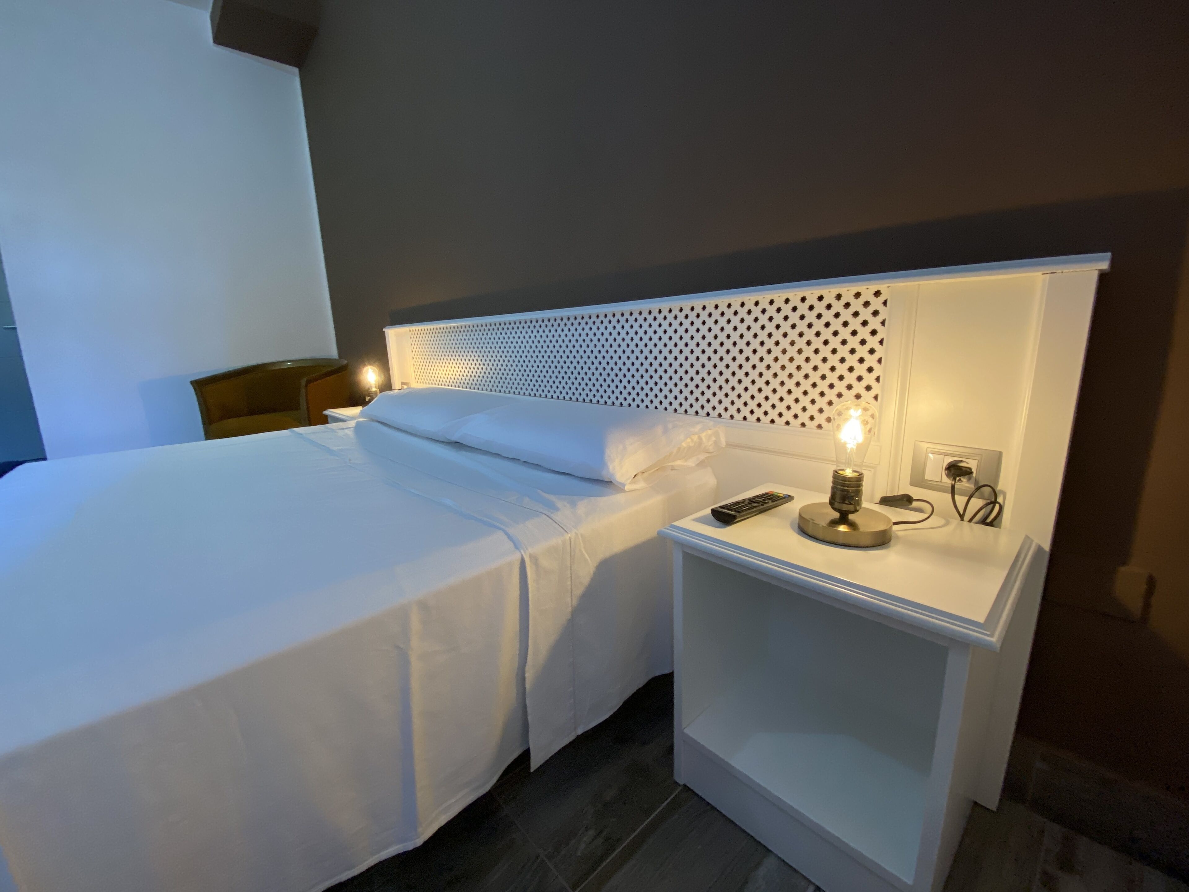 A neatly made bed with white linens, a bedside lamp, and a remote control on a white nightstand.