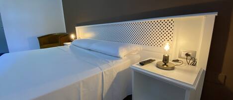 A neatly made bed with white linens, a bedside lamp, and a remote control on a white nightstand.