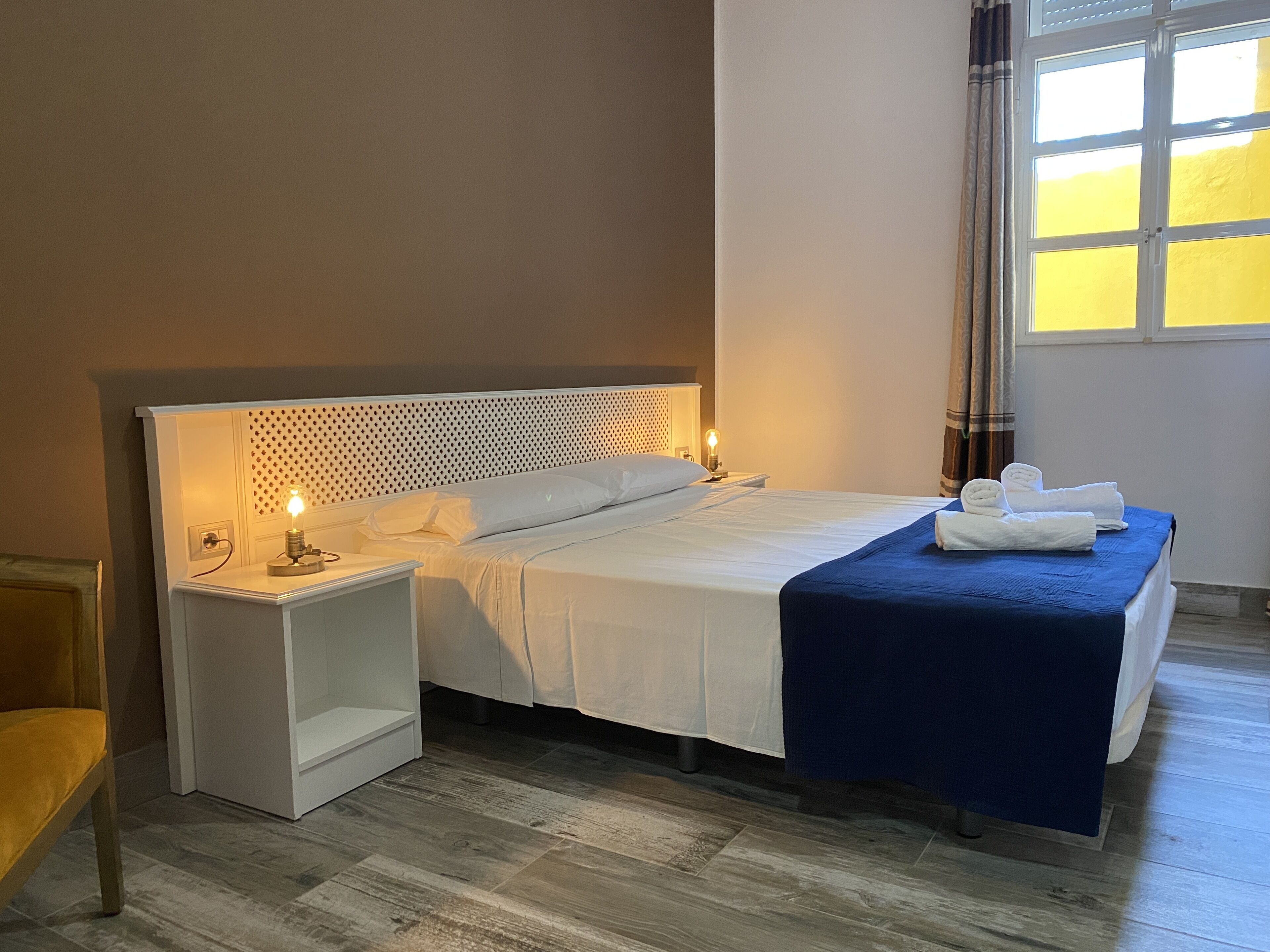 Un lit bien fait, avec des draps blancs, un plaid bleu et des serviettes soigneusement pliées, posés sur un plancher en bois.