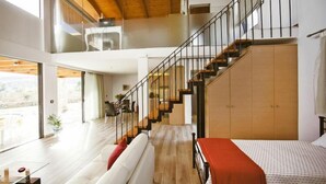Un salón amplio con una escalera que sube a un piso superior, una ventana grande y una cama con una manta roja.