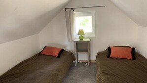 Eine kleine, einfache Unterkunft mit zwei Einzelbetten, einem Fenster mit Vorhang, einem kleinen Tisch mit Lampe und Teppichboden.