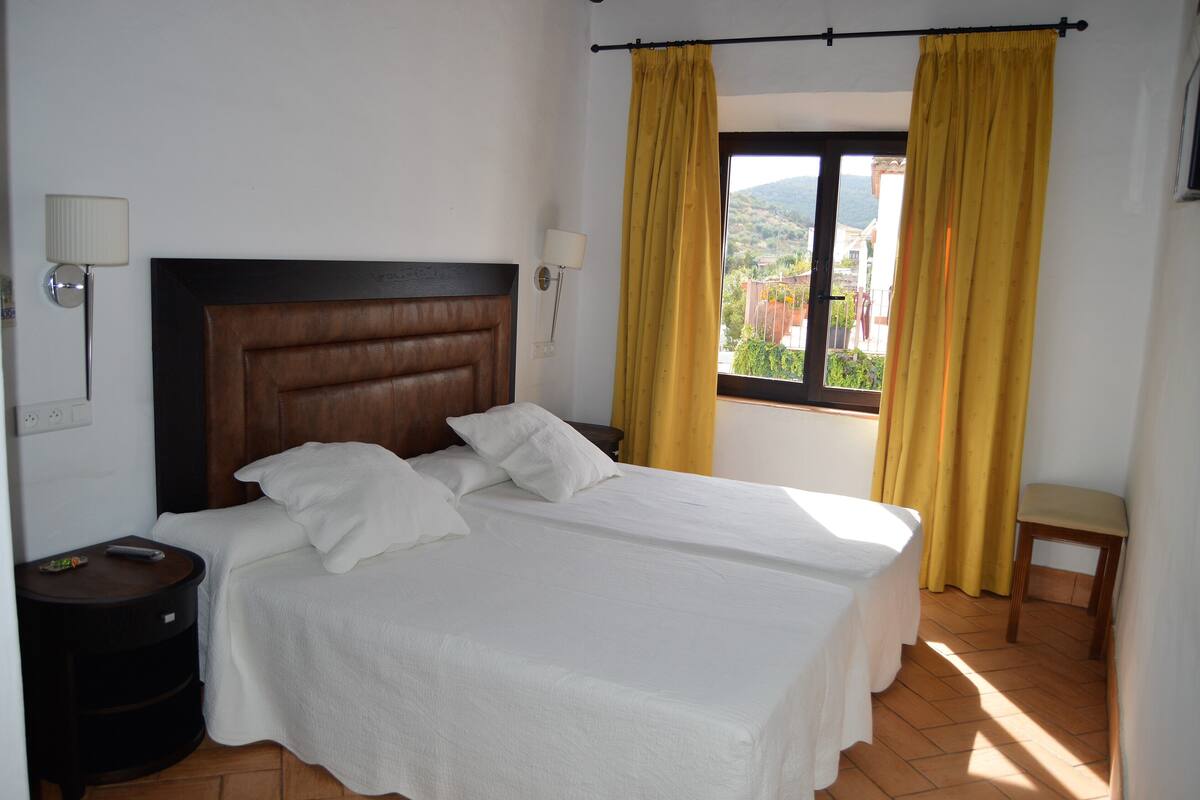 A double bed with white linens, a wooden headboard, two bedside lamps, and a window with yellow curtains.