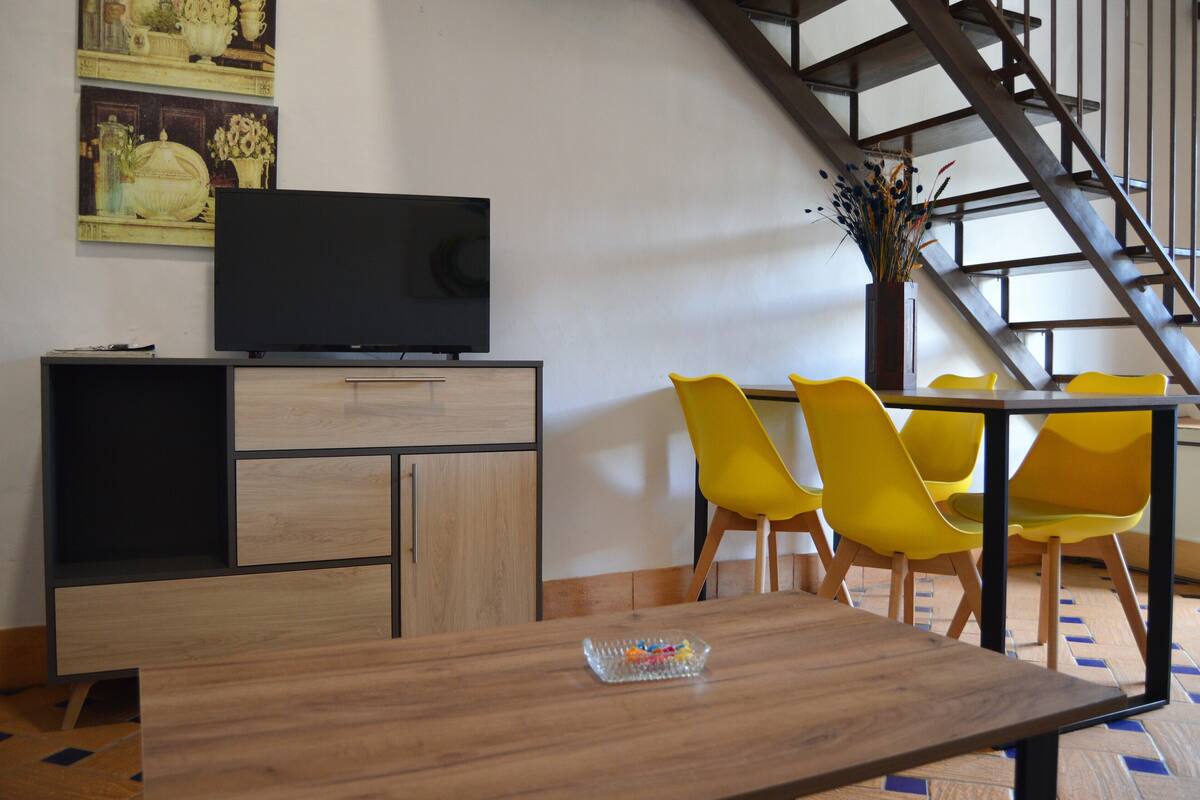 A dining area with a wooden table and yellow chairs, a television on a cabinet, and a staircase in the background.