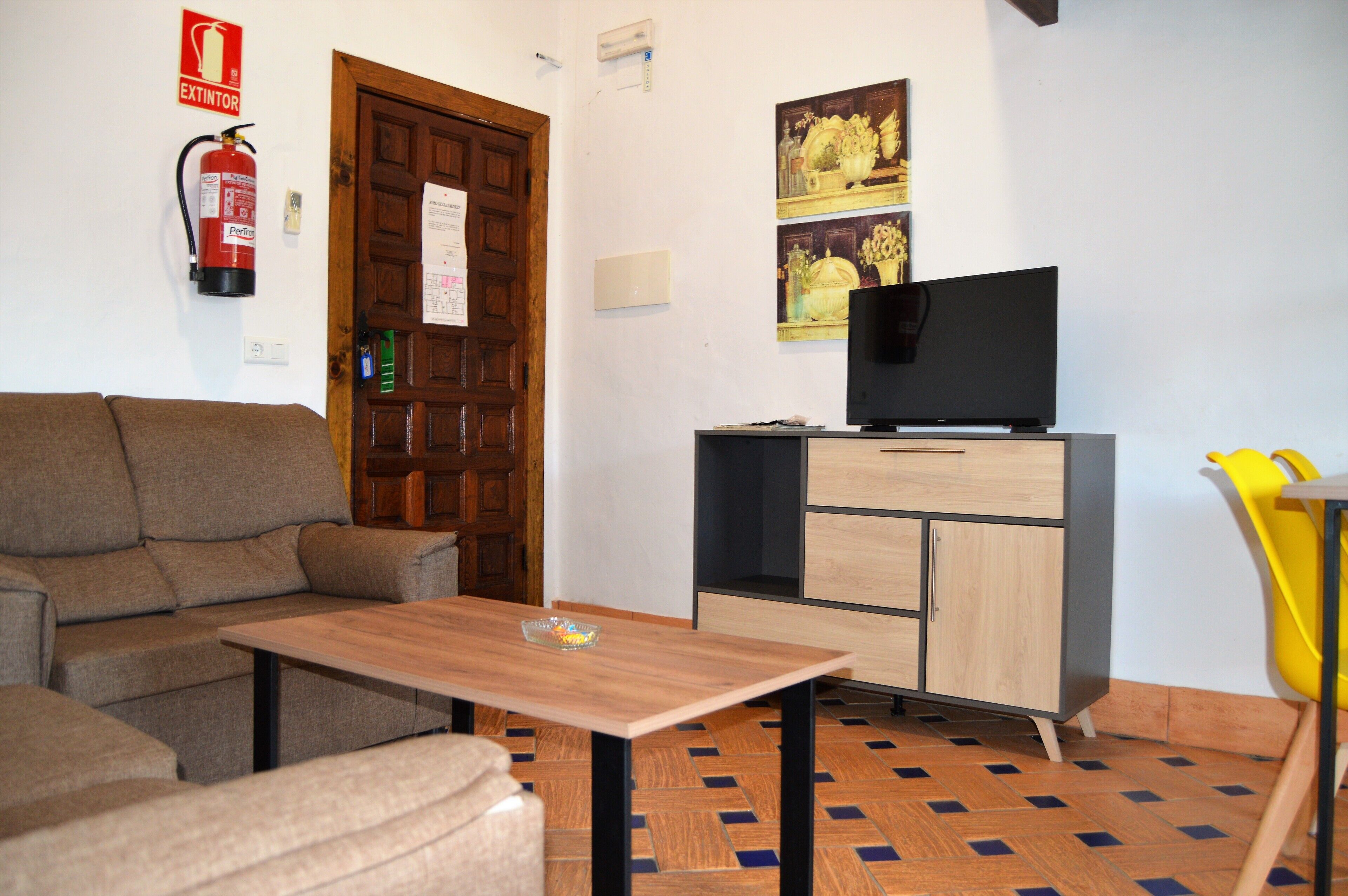 Ein Wohnzimmer mit einer Couch, einem Holztisch, einem Fernseher auf einem Schrank, einem gelben Sessel und einem Feuerlöscher an der Wand.