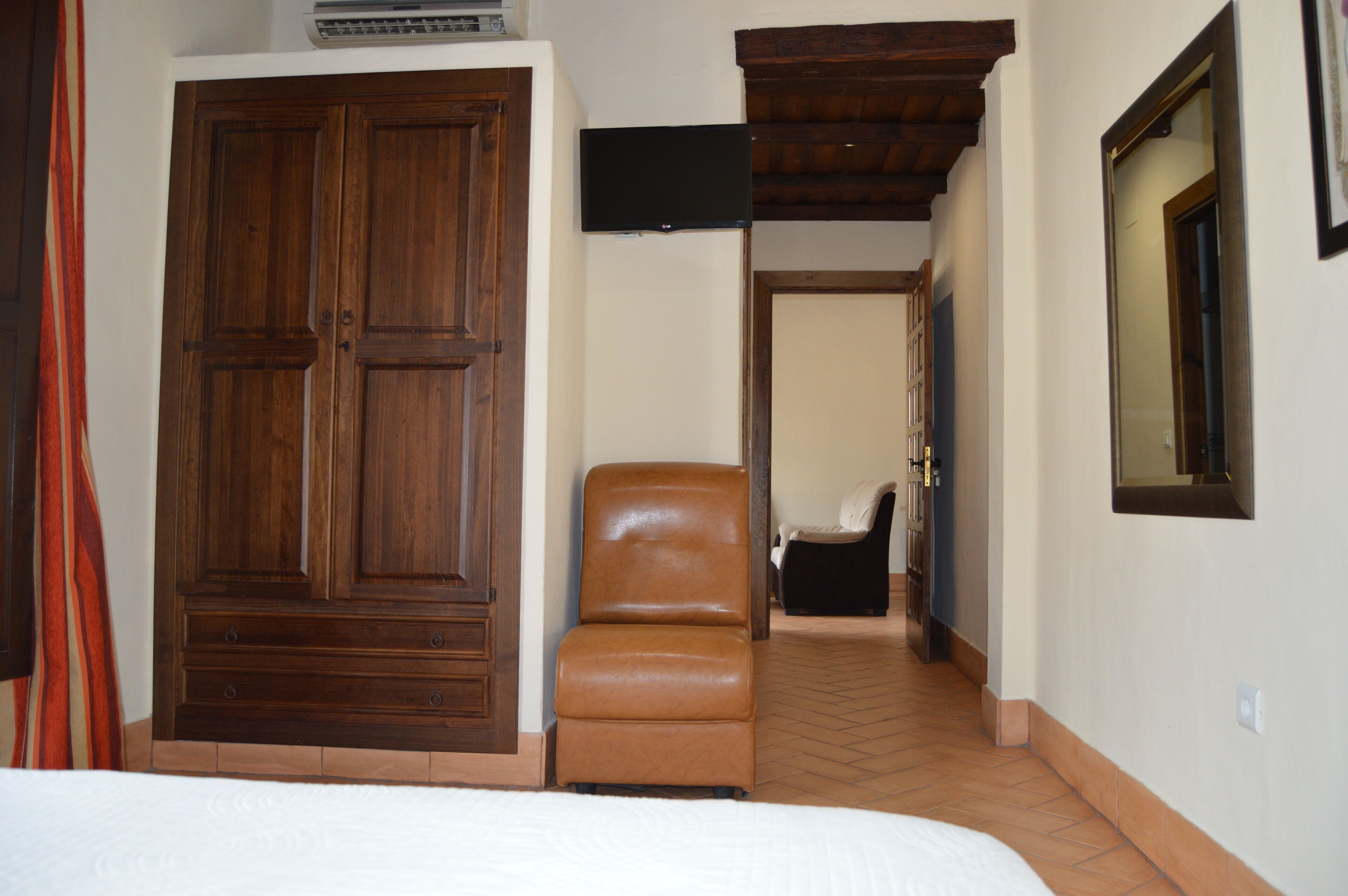 A bedroom with a large wooden wardrobe, a leather chair, a bed, and a television mounted on the wall.