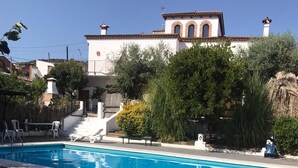 A swimming pool with a green area in the foreground, a white building with a red roof in the background, and a staircase leading to the building. - Apartamentos Turísticos Los Alisos (Aliseda)
