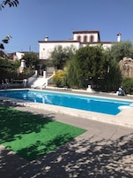 Una piscina con un área verde en primer plano, un edificio blanco con techo rojo al fondo, y una escalera que conduce al edificio.
