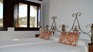 A double bed with white linens and orange patterned pillows, a wooden headboard, a nightstand with a lamp, and a window with a view of hills. - Apartamentos Turísticos Los Alisos (Aliseda)