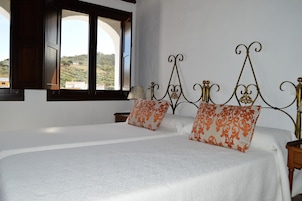 Una cama doble con sábanas blancas y almohadas anaranjadas estampadas, un cabecero de madera, una mesita de noche con una lámpara y una ventana con vistas a las colinas.