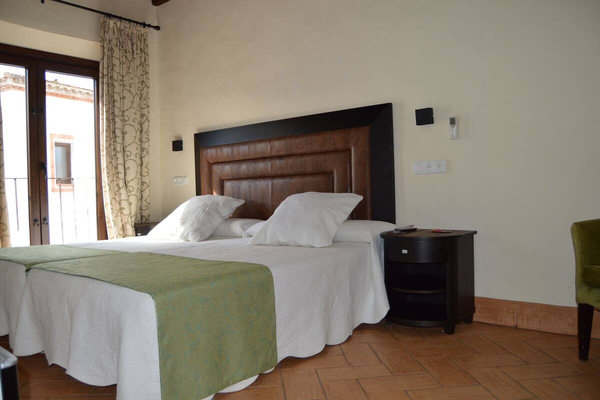 A hotel room with a large bed, a bedside table, a green chair, and a view of the outside.