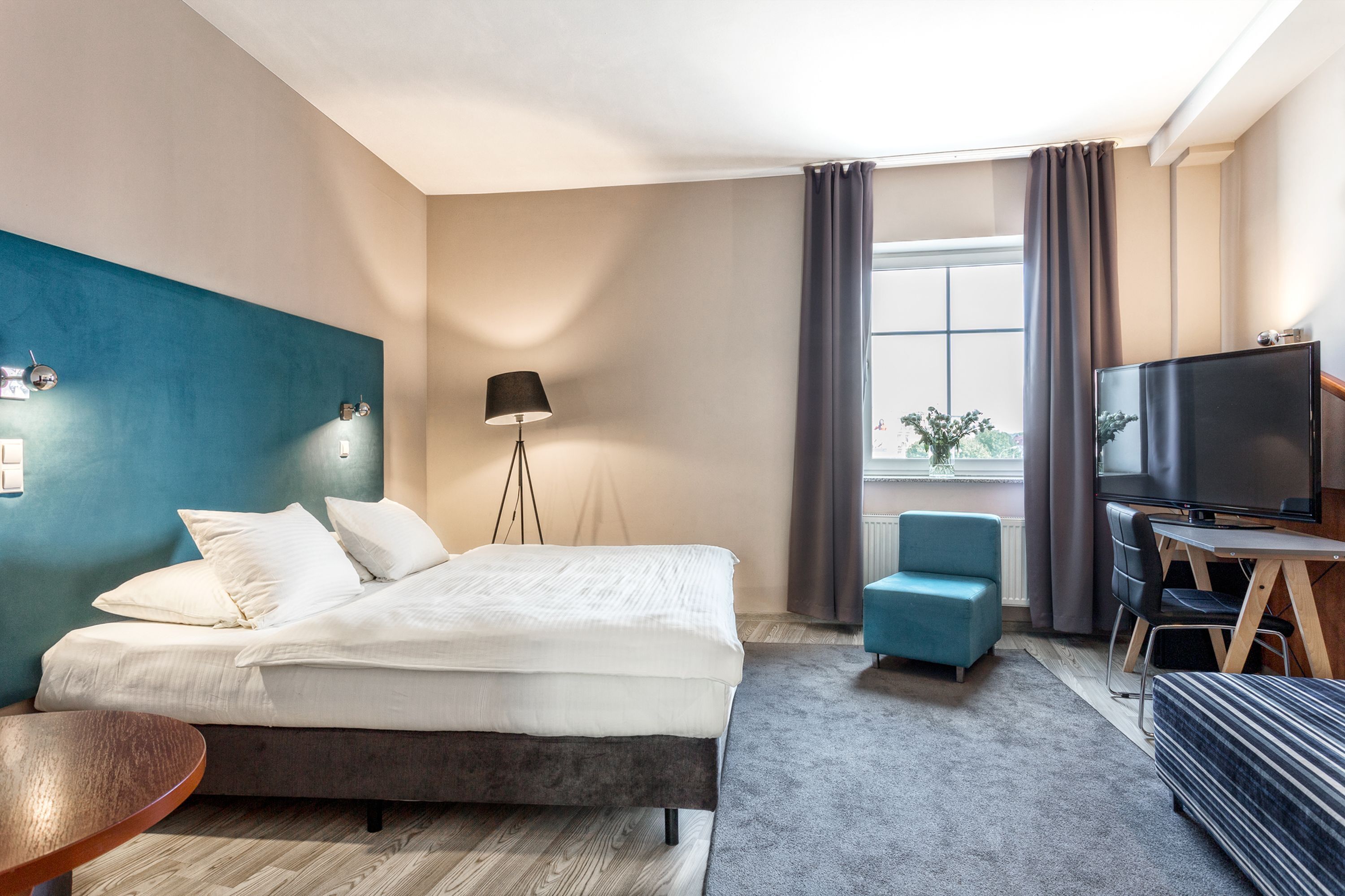 Een moderne hotelkamer met een groot bed, een bureau met televisie, een blauwe fauteuil en een vloerlamp.