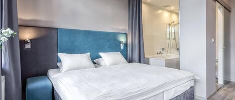 Habitación de hotel moderna con una cama grande, una mesita de noche, un espejo y un baño visible a través de una puerta abierta.