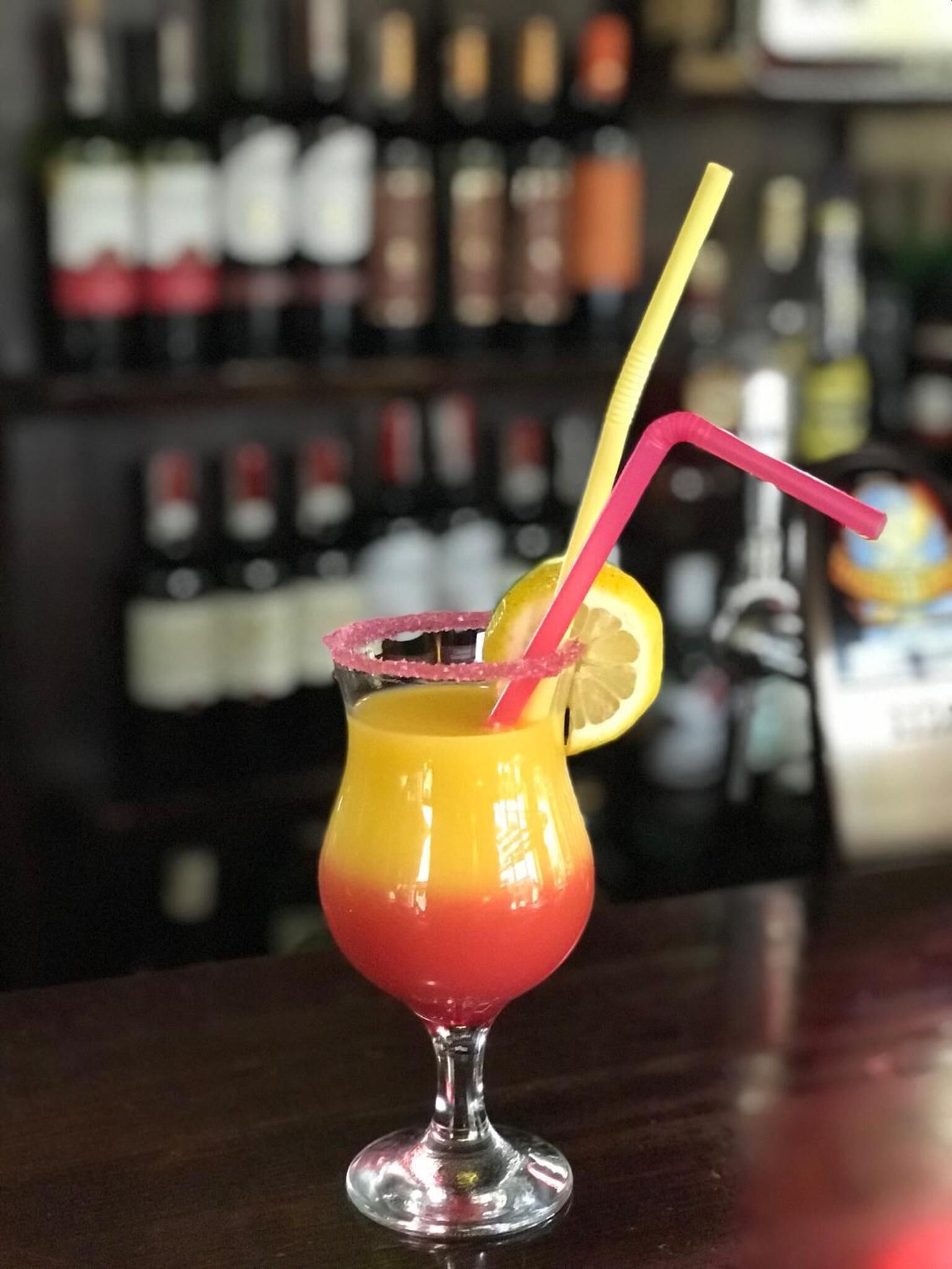 Um cocktail num copo com uma fatia de limão e duas canudos, apoiado numa bancada de bar com garrafas ao fundo.