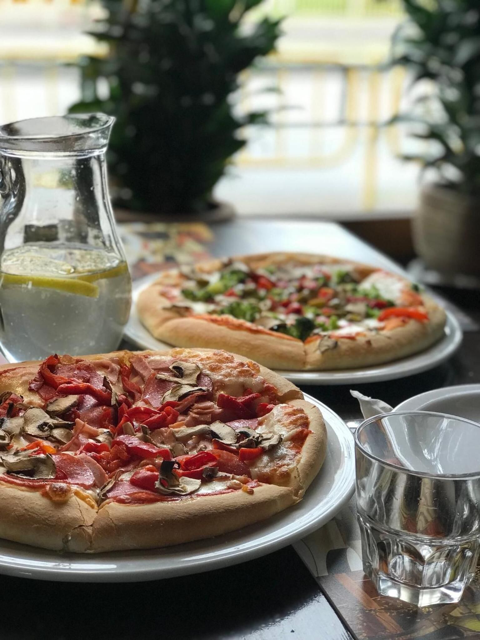 Two pizzas on plates with a pitcher of water and a glass on a table.