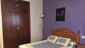 Une chambre avec une armoire en bois, un lit avec une literie à motifs, une lampe de sol et un tableau représentant des fleurs accroché au mur.