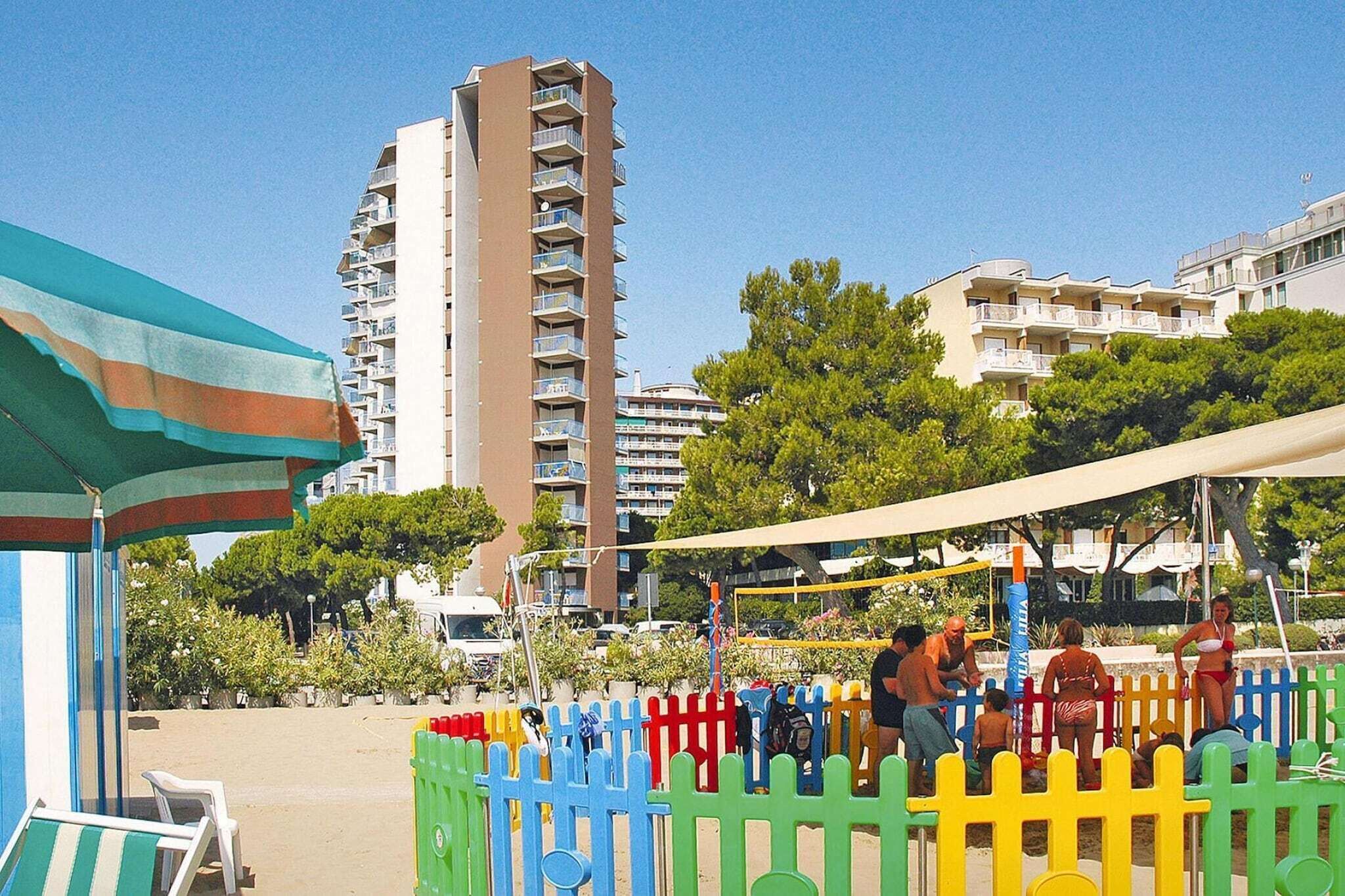 A beach area with a tall building in the background, a colorful fence, and a beach umbrella.