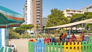 A beach area with a tall building in the background, a colorful fence, and a beach umbrella. - Apartments Bianco Nero, Lignano Sabbiadoro (Lignano Sabbiadoro)