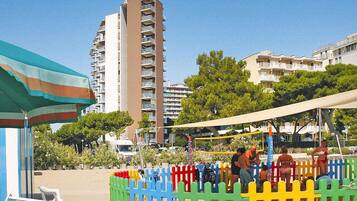 A beach area with a tall building in the background, a colorful fence, and a beach umbrella.