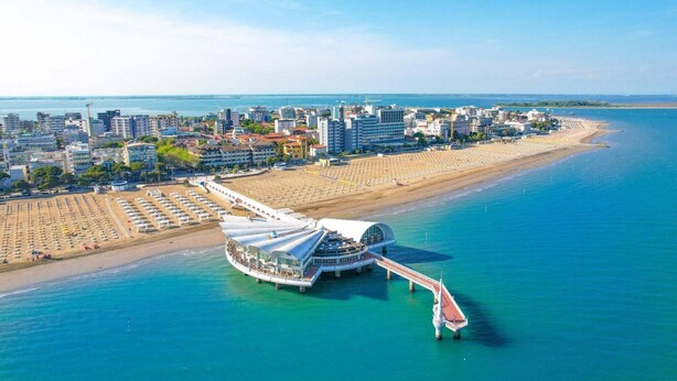 An aerial view of a coastal city with a prominent waterfront promenade, a large white building with a curved roof, and a beach with rows of umbrellas. - Apartments Bianco Nero, Lignano Sabbiadoro (Lignano Sabbiadoro)