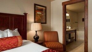 Habitación de hotel con cama, una silla, lámpara, un cuadro y baño visible a través de una puerta abierta.