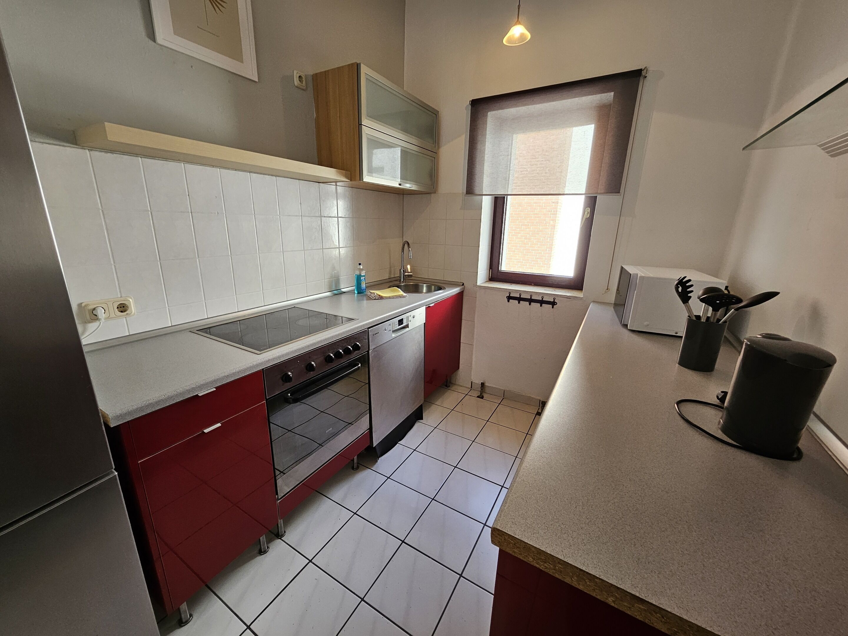 Een kleine keuken met een kookplaat, oven en vaatwasser, met rode kastjes en witte tegels.