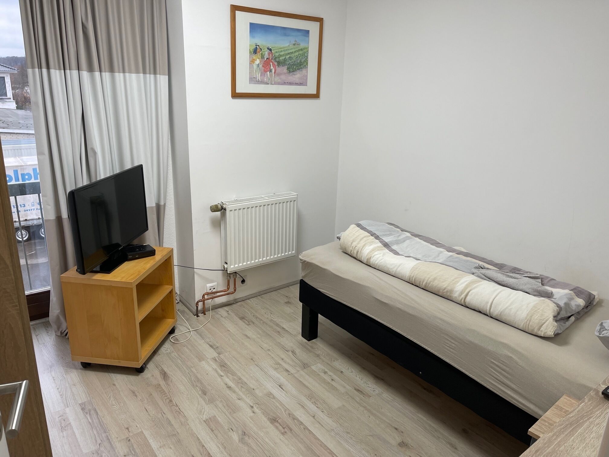 Una camera da letto con un letto, una TV su un mobile in legno, un termosifone e una finestra con tende.