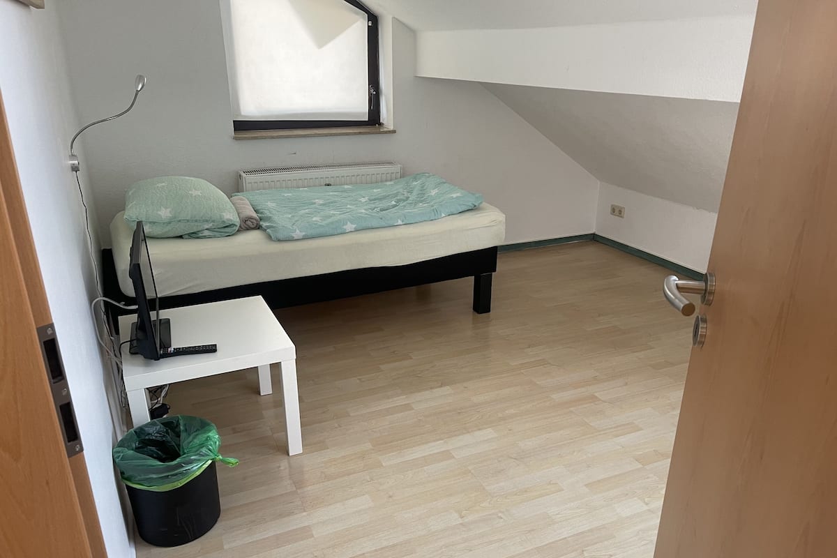 Una habitación pequeña con cama individual, techo inclinado, ventana, radiador, escritorio y un cubo de basura.