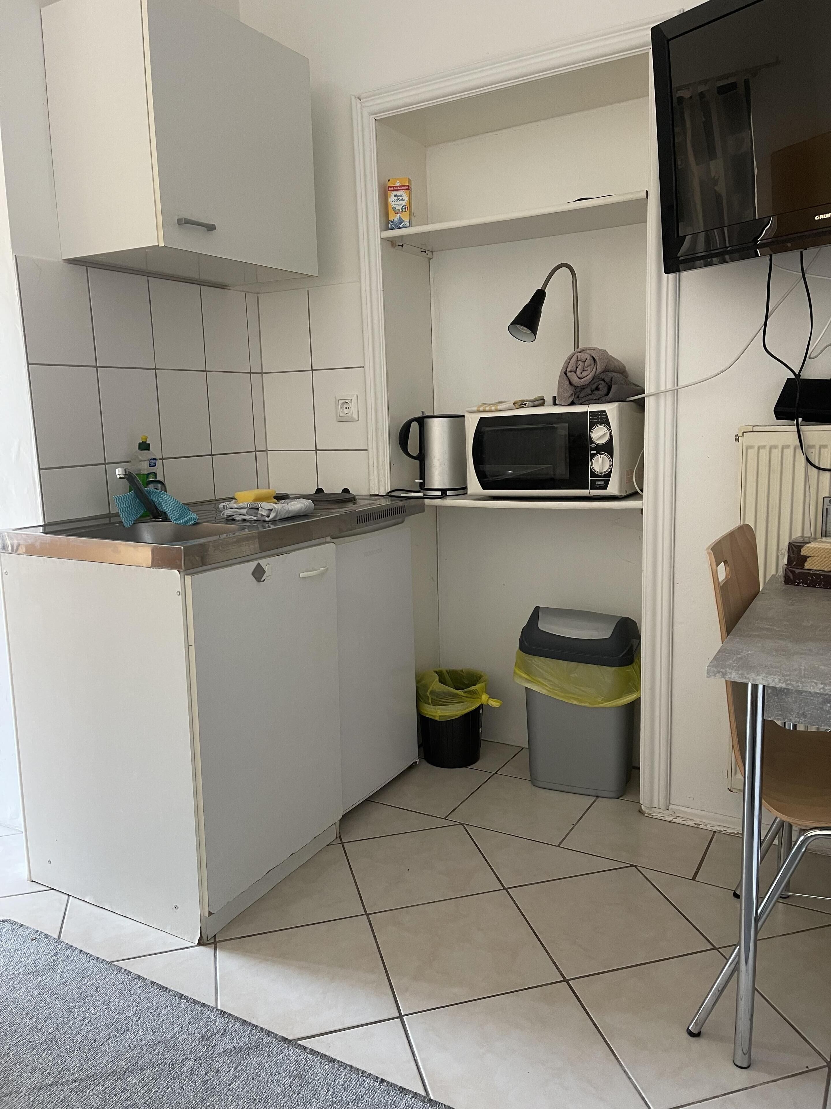 Una cocina compacta con armarios blancos, un microondas, un hervidor y un estante pequeño con una caja. Hay dos cubos de basura y una silla en la mesa.