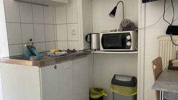 Una cocina compacta con armarios blancos, un microondas, un hervidor y un estante pequeño con una caja. Hay dos cubos de basura y una silla en la mesa.