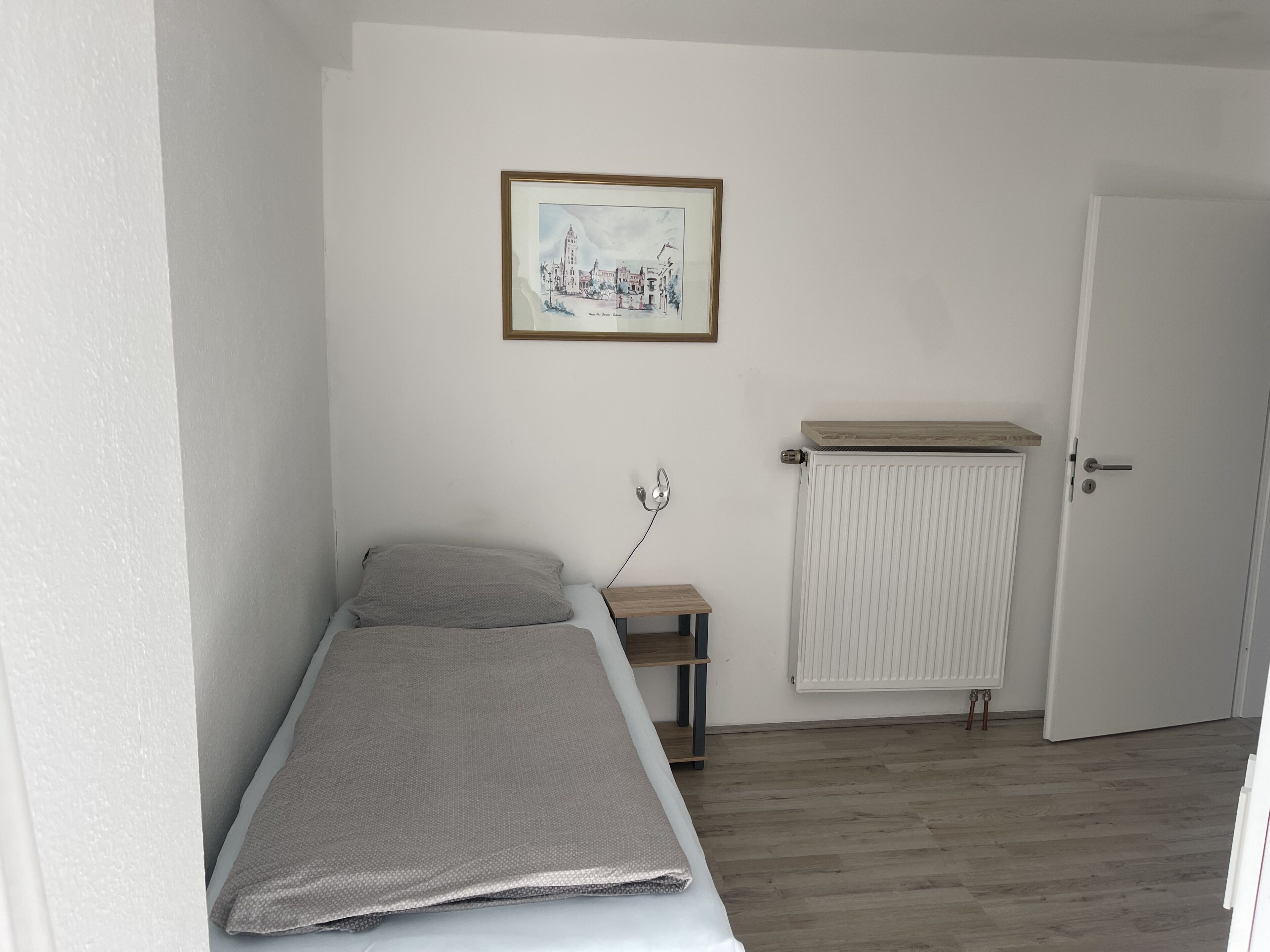 A small, single-bed room with a wooden floor, a radiator, a bedside table, and a framed picture on the wall.