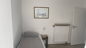 A small, single-bed room with a wooden floor, a radiator, a bedside table, and a framed picture on the wall.