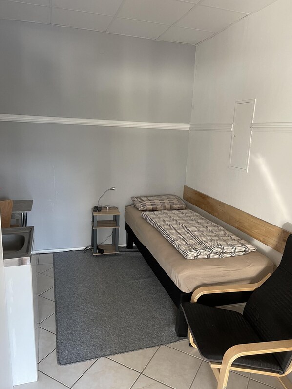 A small, single-bed room with a grey rug, a wooden chair, a desk with a lamp, and a small shelf.