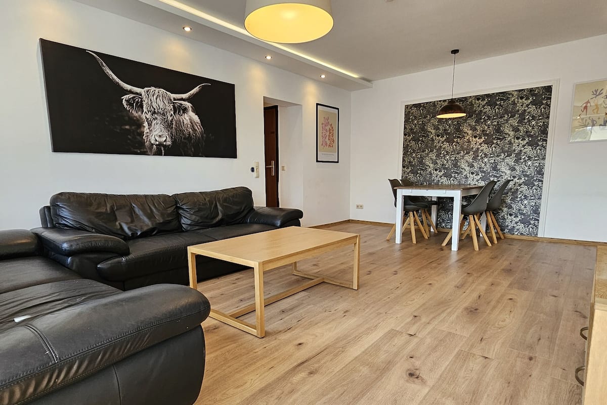 Una sala moderna con un sofá de cuero negro, una mesa de centro de madera, una mesa de comedor con sillas y un cuadro abstracto blanco y negro en la pared.