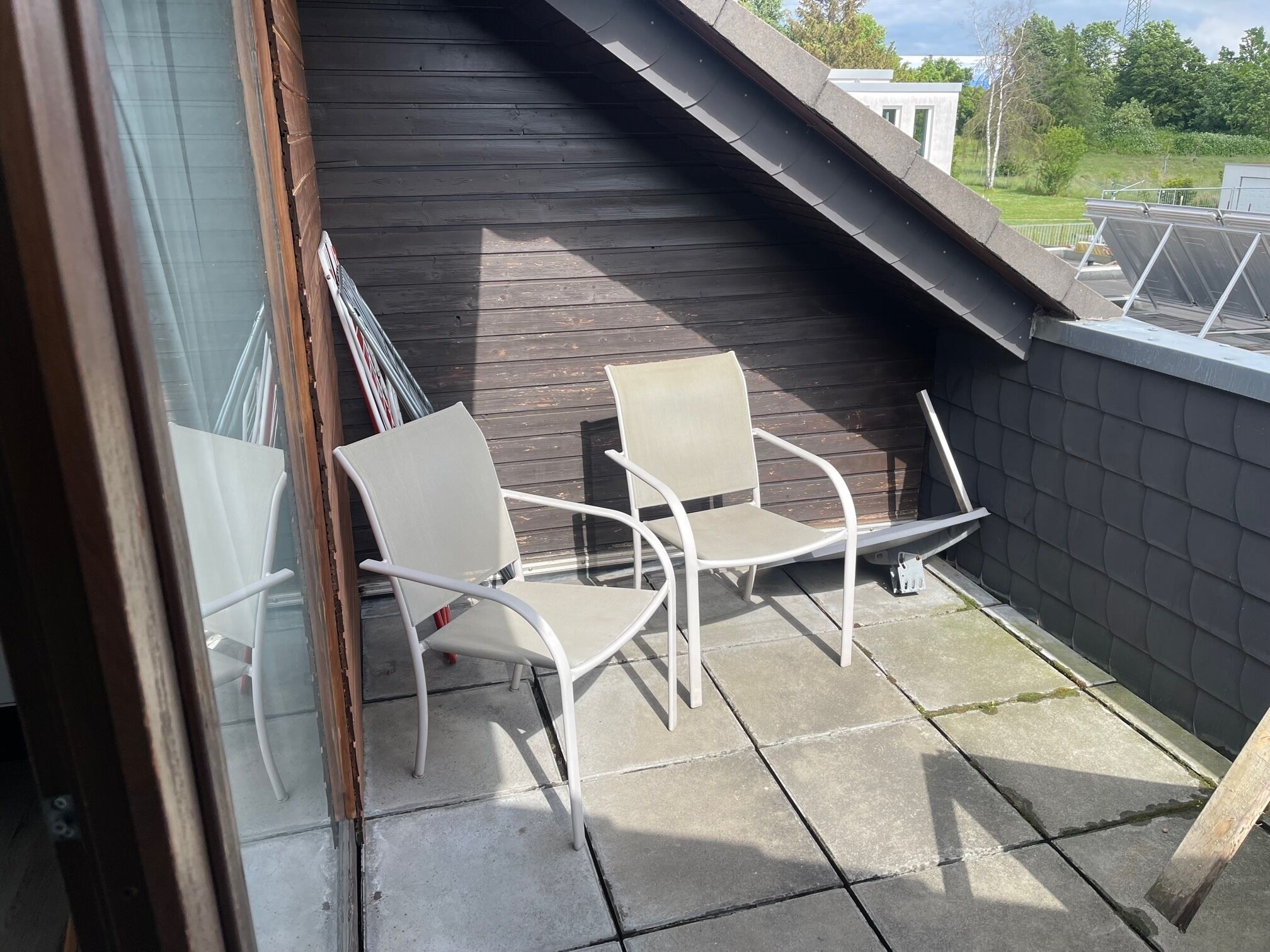 A balcony with two white chairs, a wooden wall, and a view of solar panels and greenery.