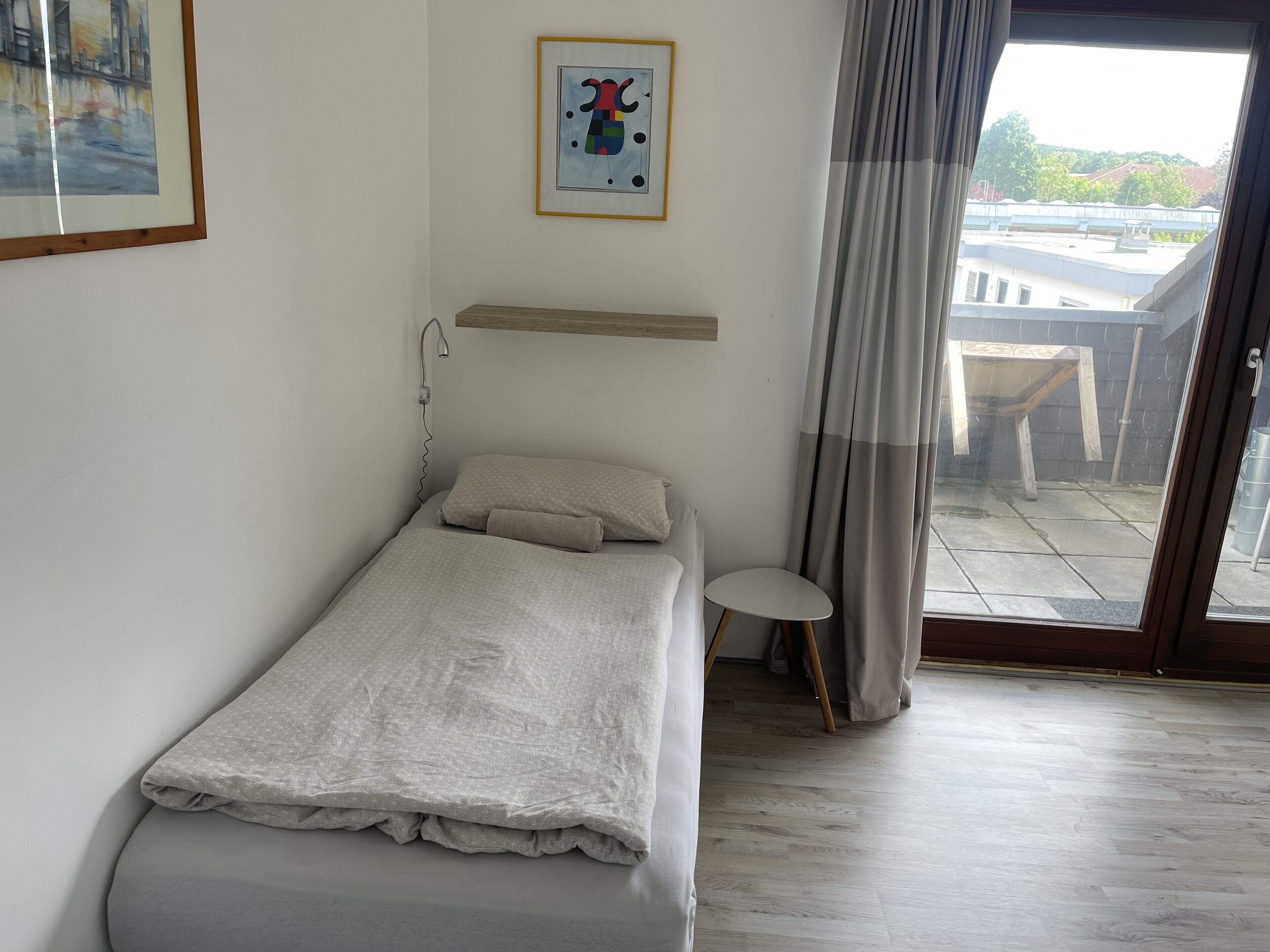 Een enkel bed met houten hoofdbord, een klein nachtkastje en een raam met gordijnen.