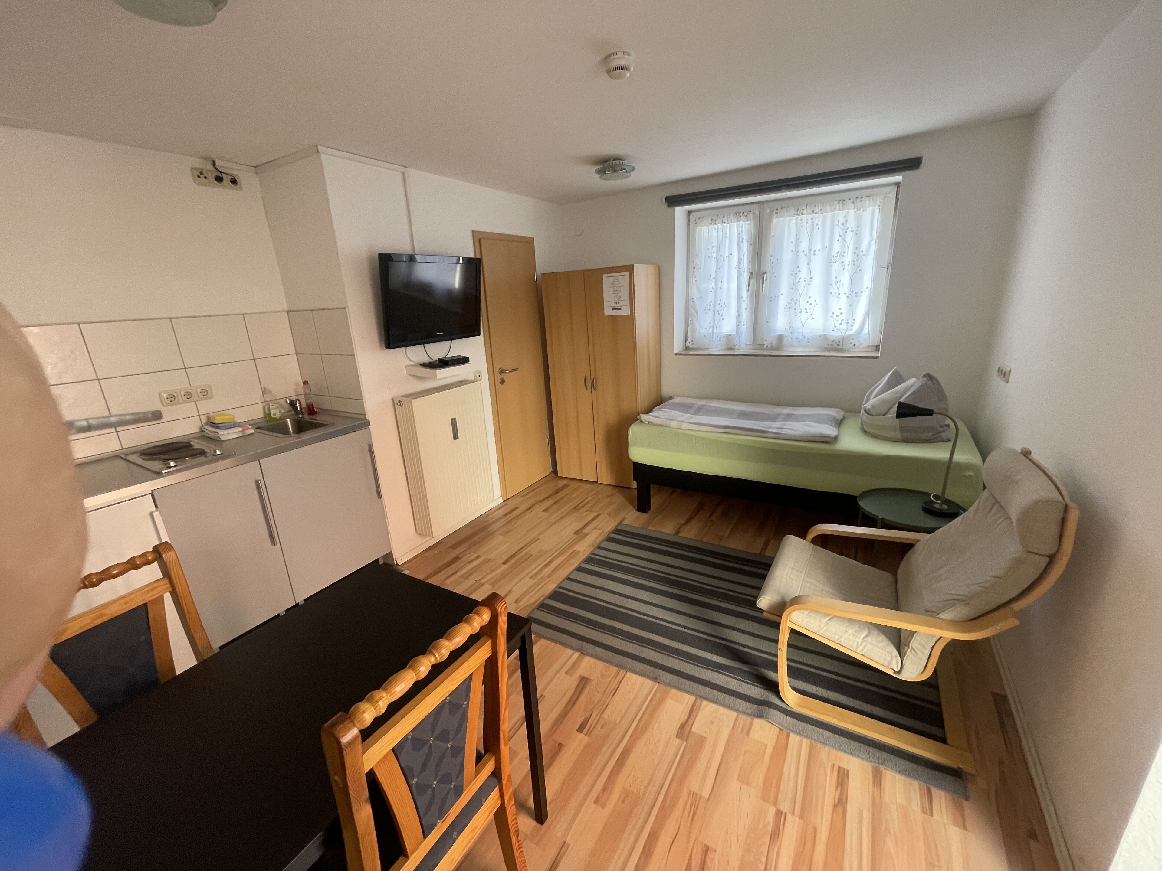A compact room with a kitchenette, a bed, a TV, and a window with curtains.