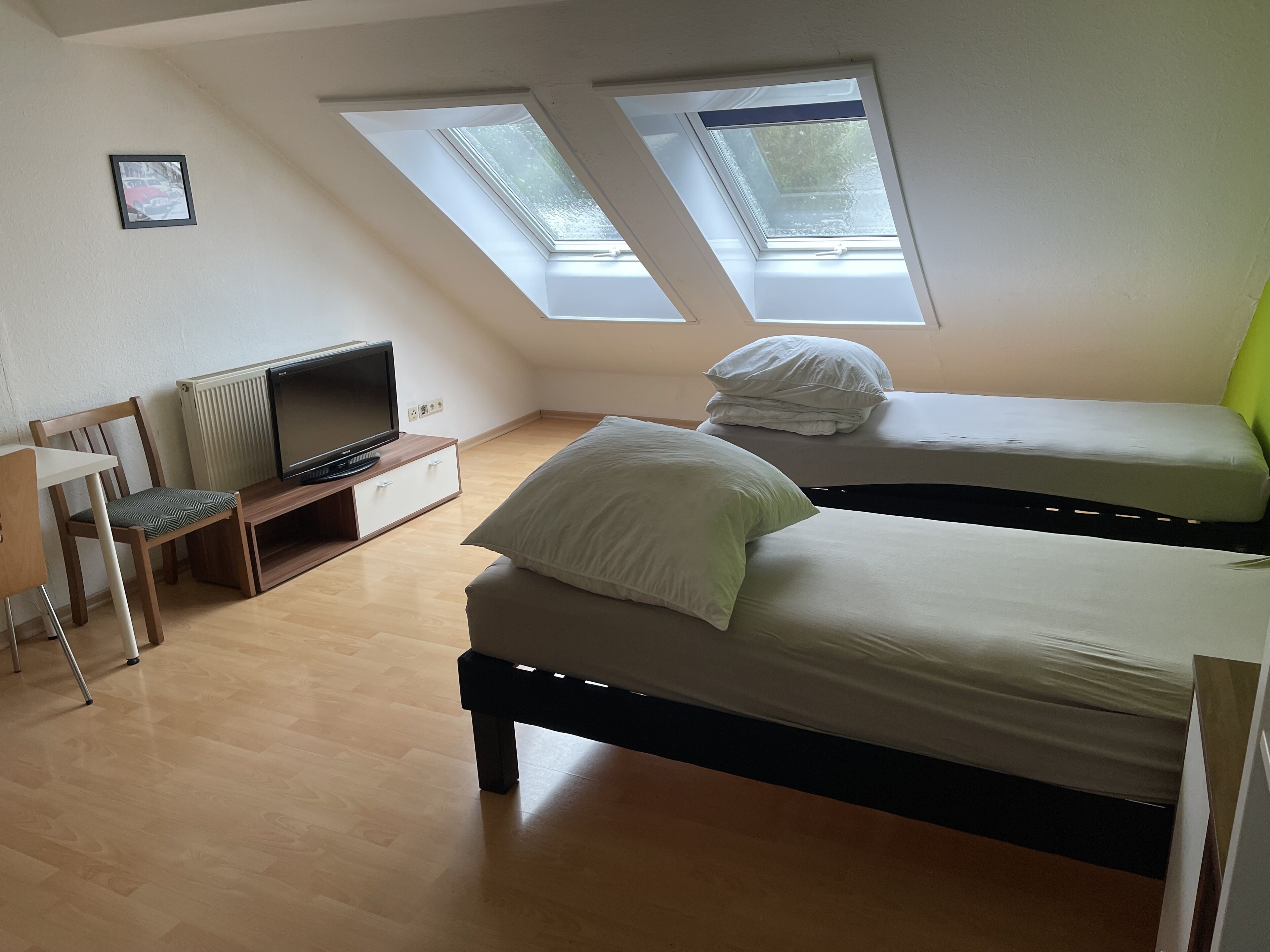 A small, well-lit room with two beds, a TV, and a desk.