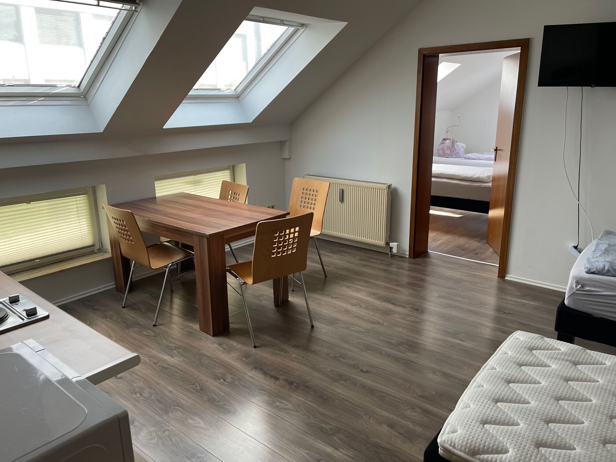 Ein modernes Zimmer mit einem Holzesstisch, vier Stühlen, einem Bett, einem Fernseher und Dachfenstern.