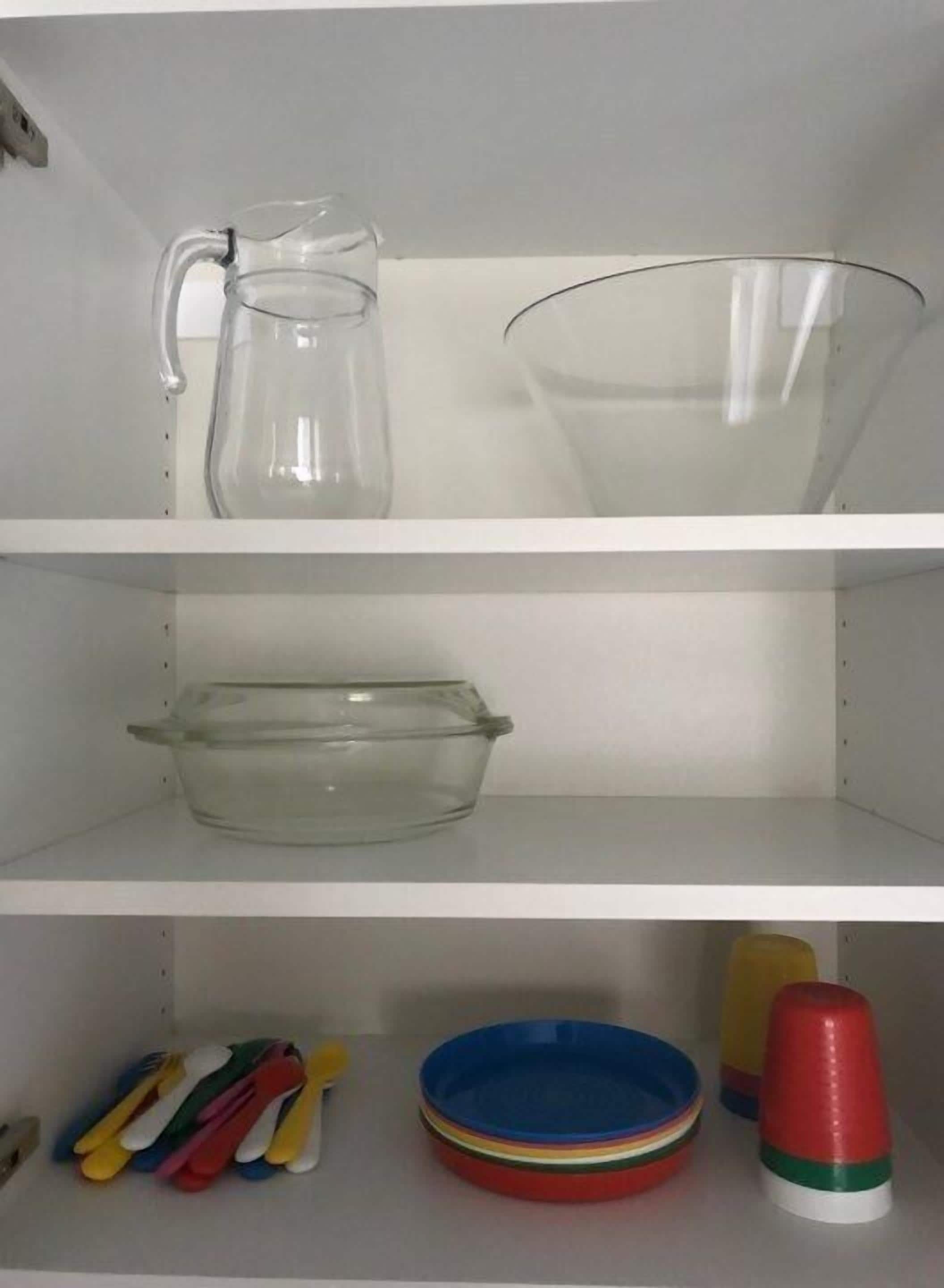 A kitchen shelf with a pitcher, a glass bowl, a glass baking dish, and colorful plastic utensils.