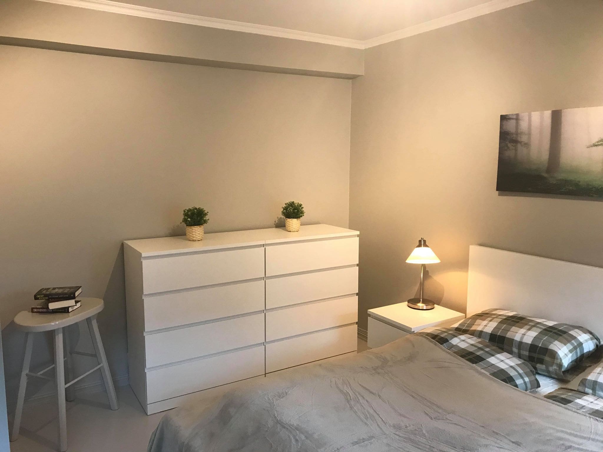 A bedroom with a bed, a dresser, a nightstand, a lamp, and a small stool.